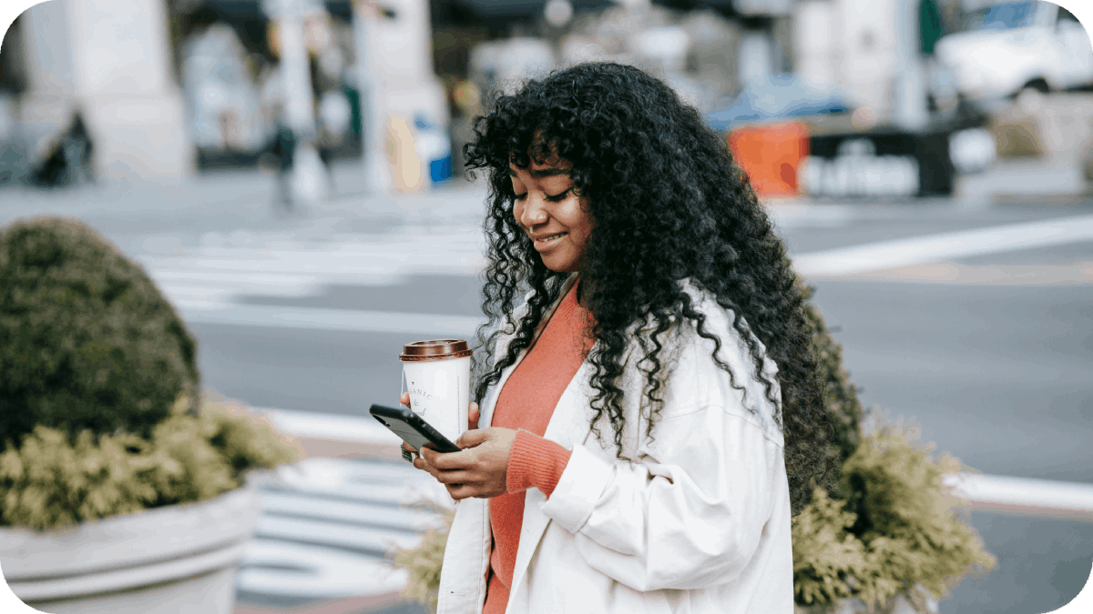 A black women in the city looking at her phone and holding the coffee cup in the other hand