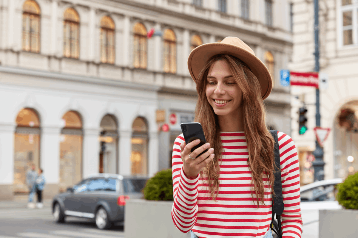 Smiling young European woman enjoys recreation time, watches her slick app, wearing a stylish headgear and striped sweater