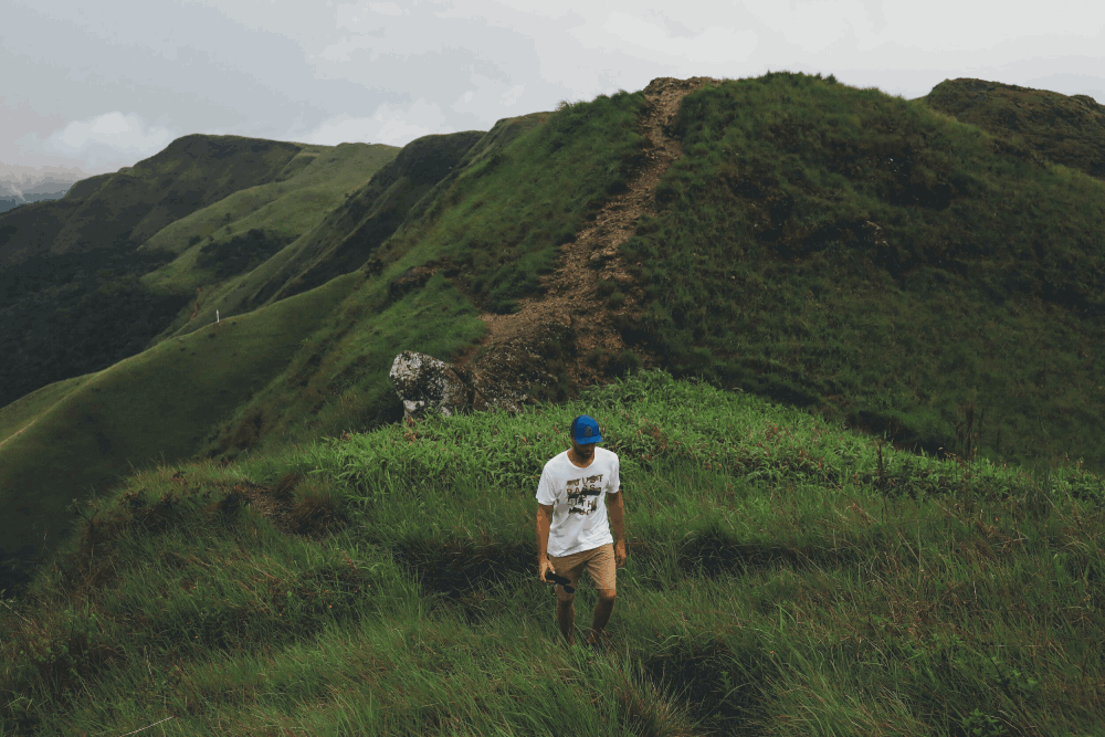 A hiker in a white T-shirt and blue cap walks up a green mountain trail surrounded by misty hills.