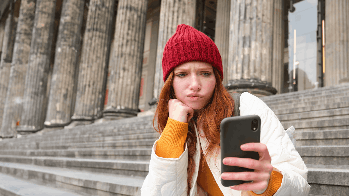 Portrait of young redhead woman with complicated face sits on street stairs in red hat holds a phone with roaming issues