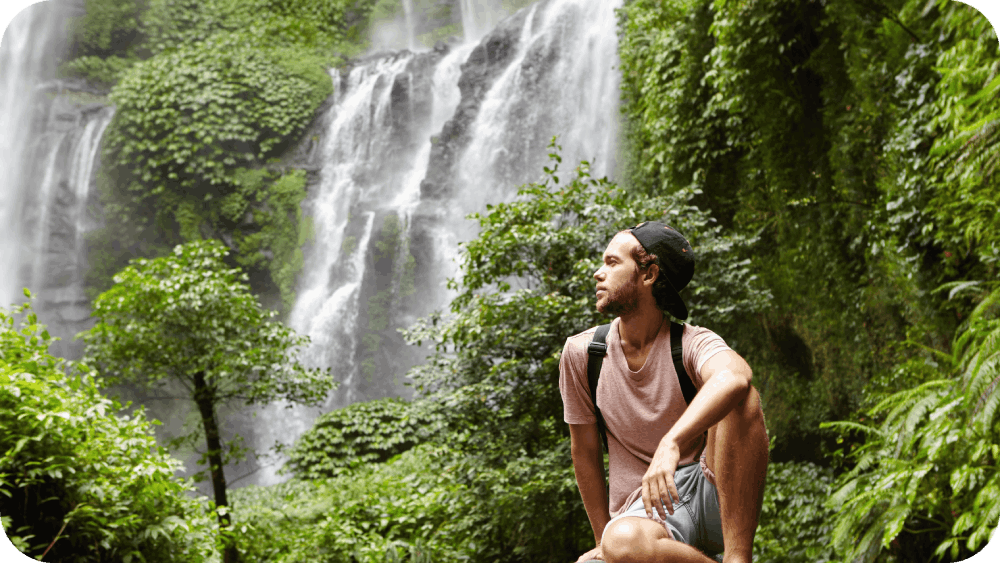 young caucasian barefooted male tourist with backpack sitting-rock surrounded by rainforest admiring gorgeous view with waterfall