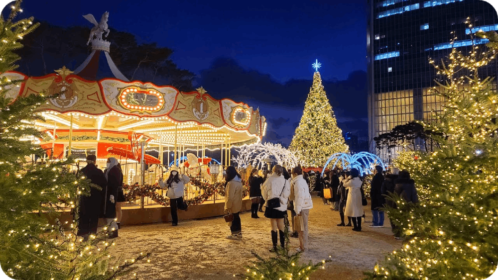 Festive holiday scene featuring a beautifully illuminated carousel, a towering Christmas tree, and people enjoying a winter wonderland atmosphere at night.